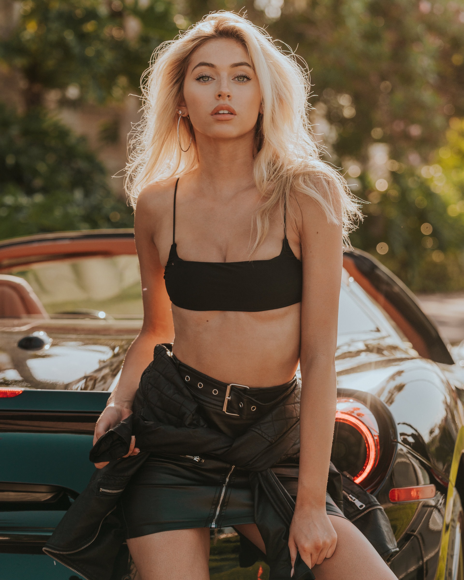 Blonde model in black bralette and leather skirt leans against a sports car outdoors.