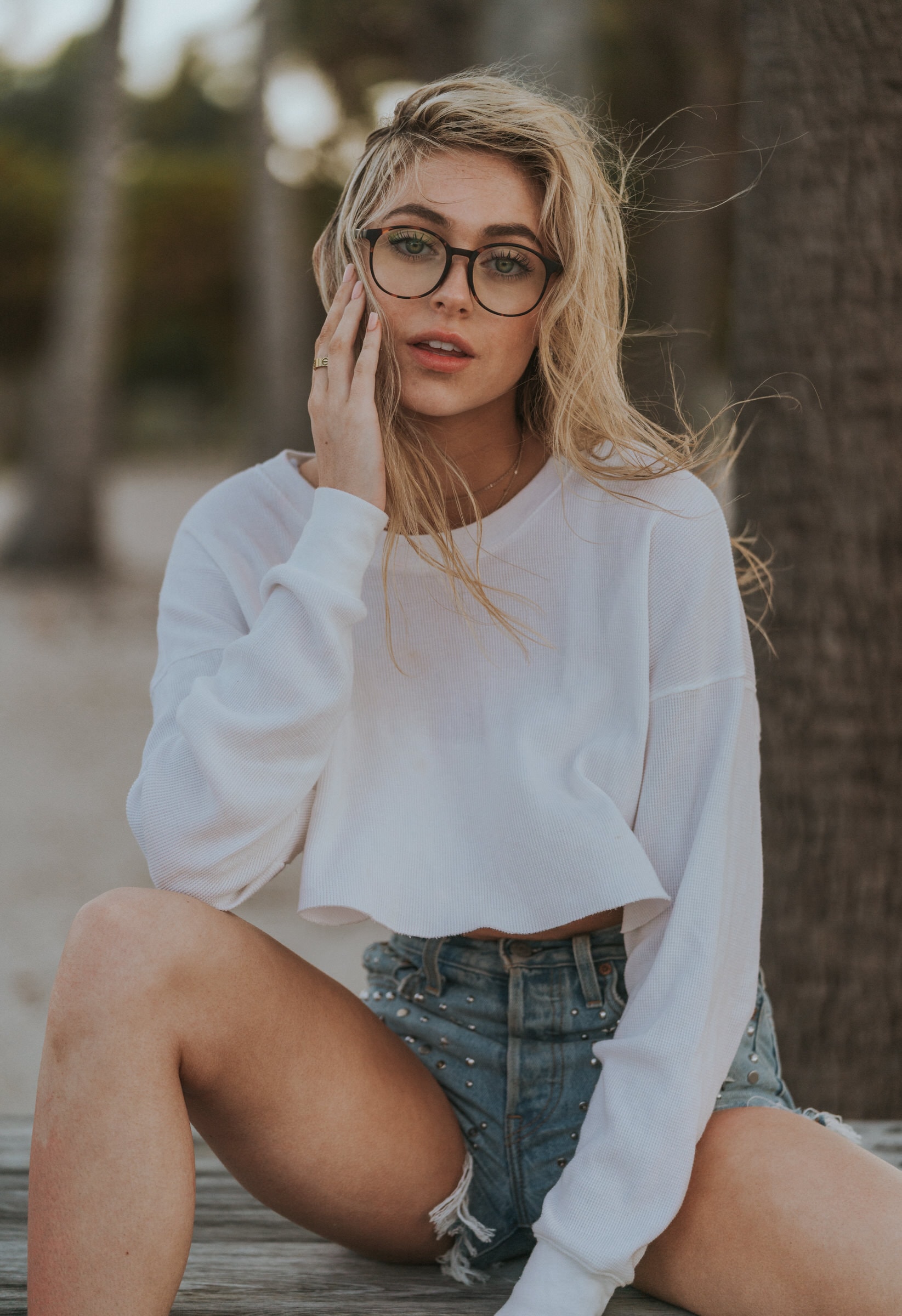 Portrait of a woman in glasses and denim shorts sitting outdoors near palm trees.
