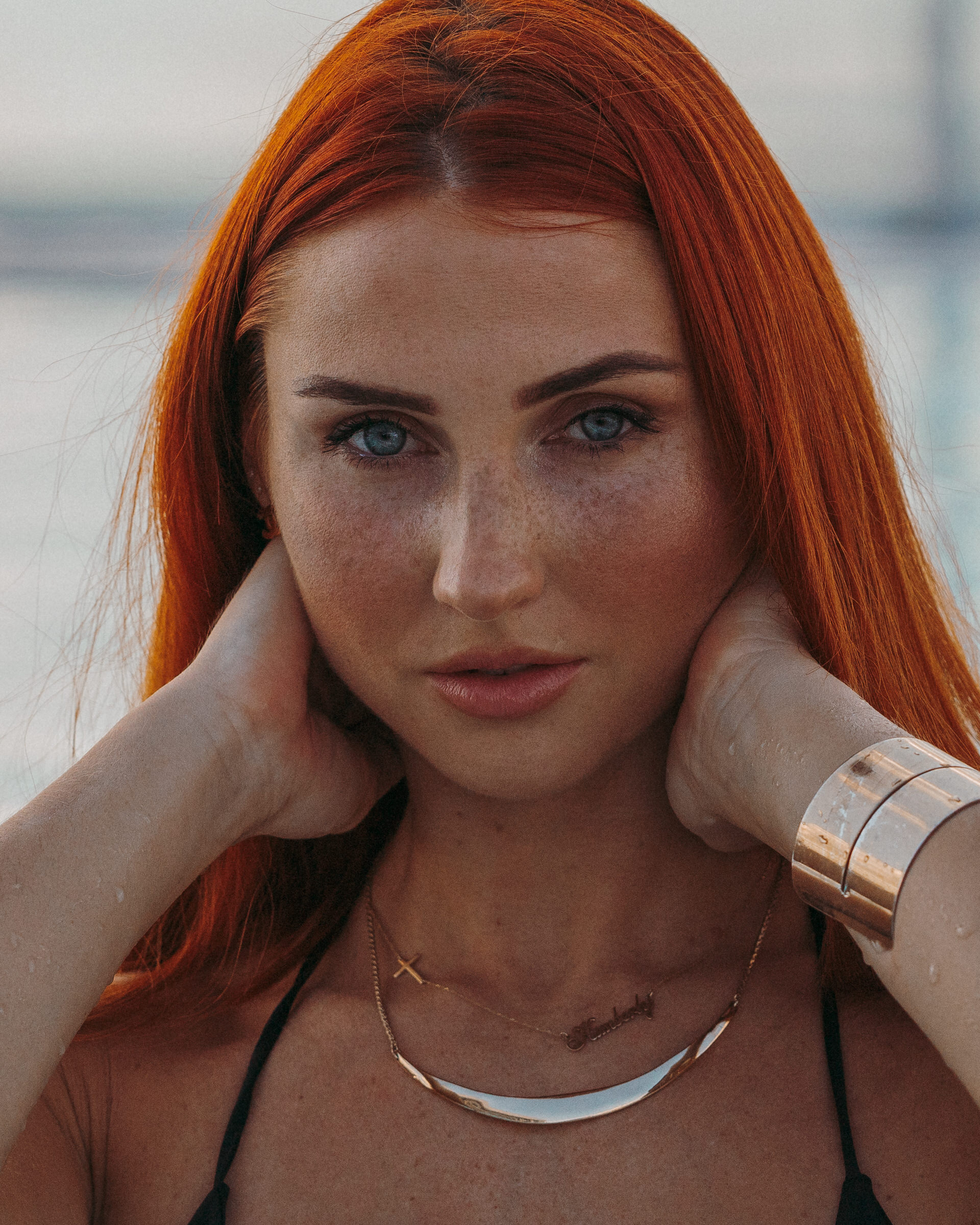 Close-up beach portrait of a red-haired woman wearing layered necklaces and a metal cuff bracelet.