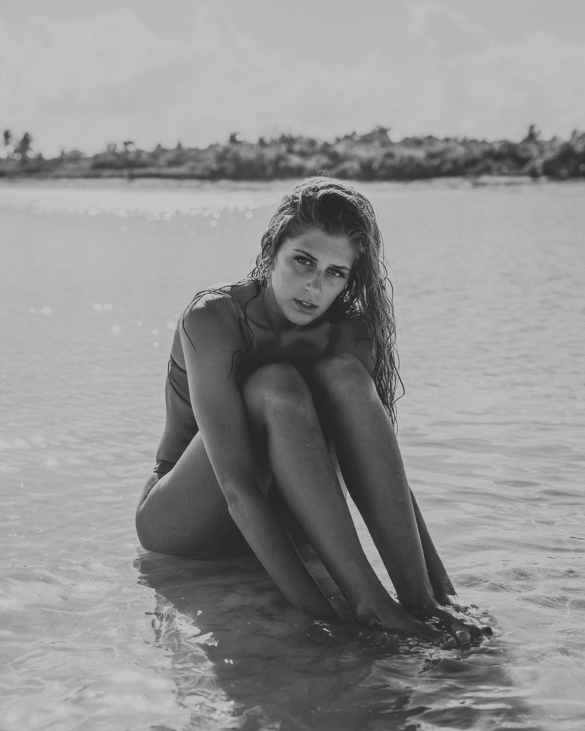 Black and white portrait of a woman sitting in shallow water, knees drawn up.
