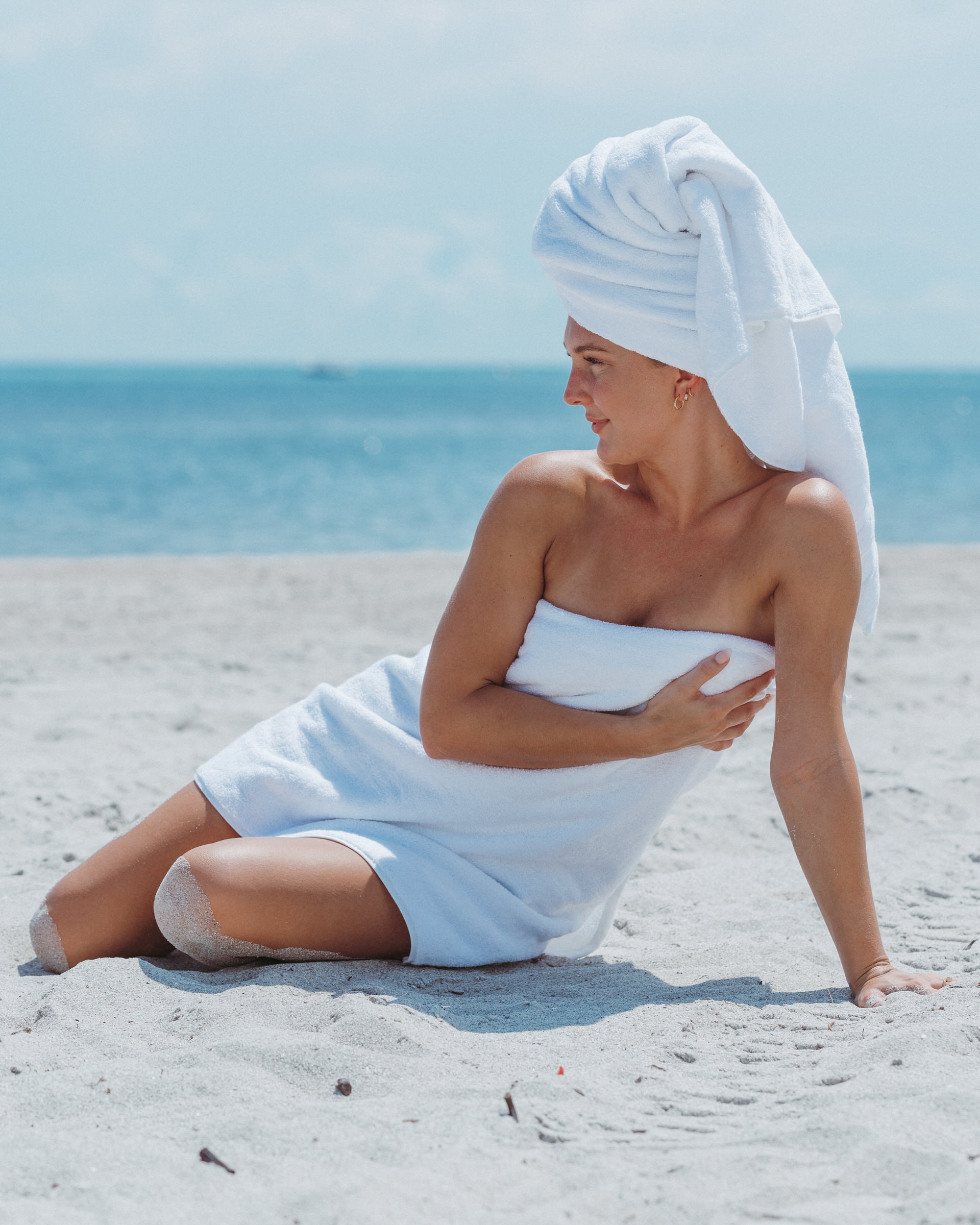 Woman wrapped in white towels sits on the sand, ocean stretching behind her.