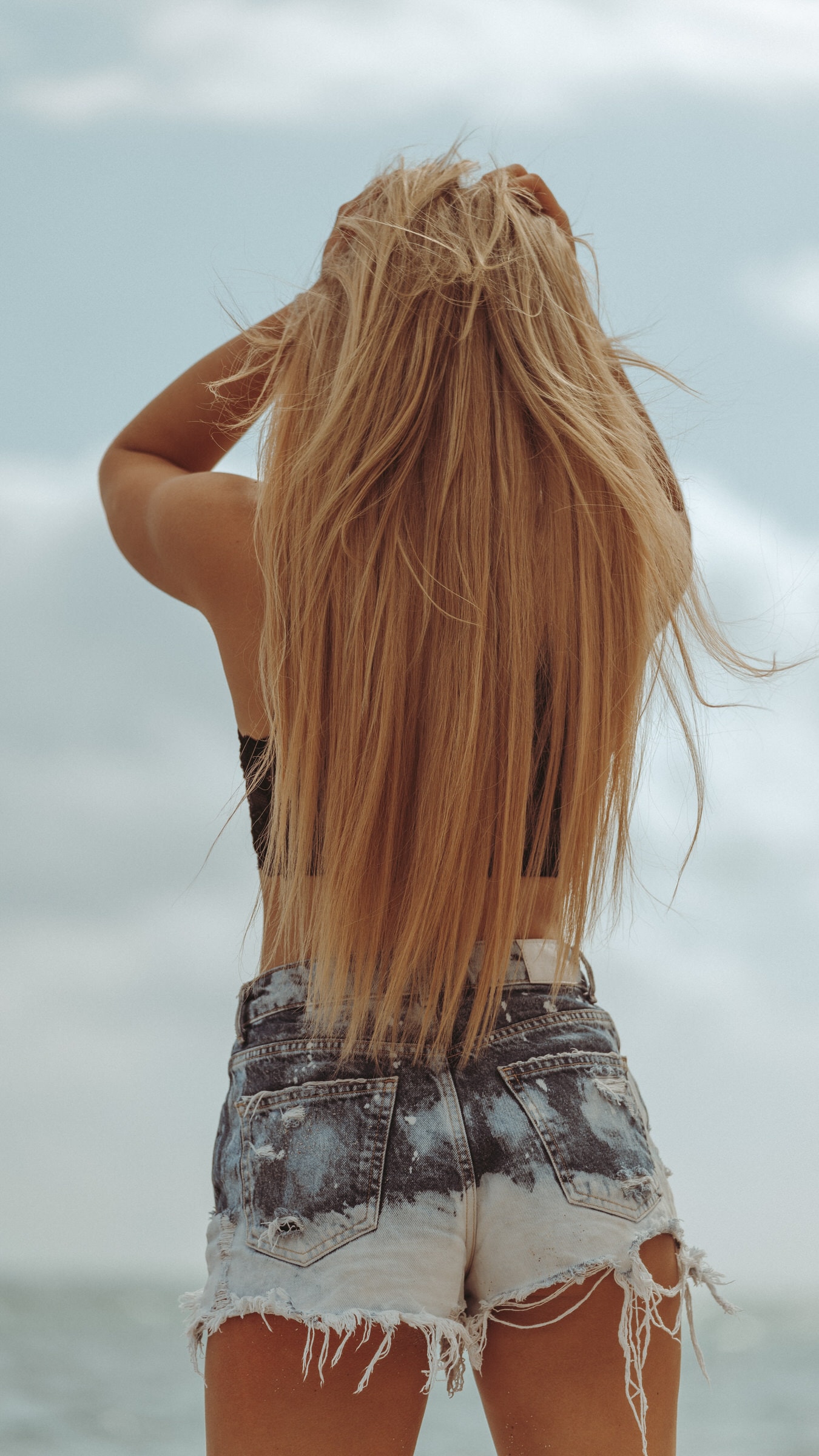 Woman with long blonde hair in denim shorts faces the ocean, seen from behind.