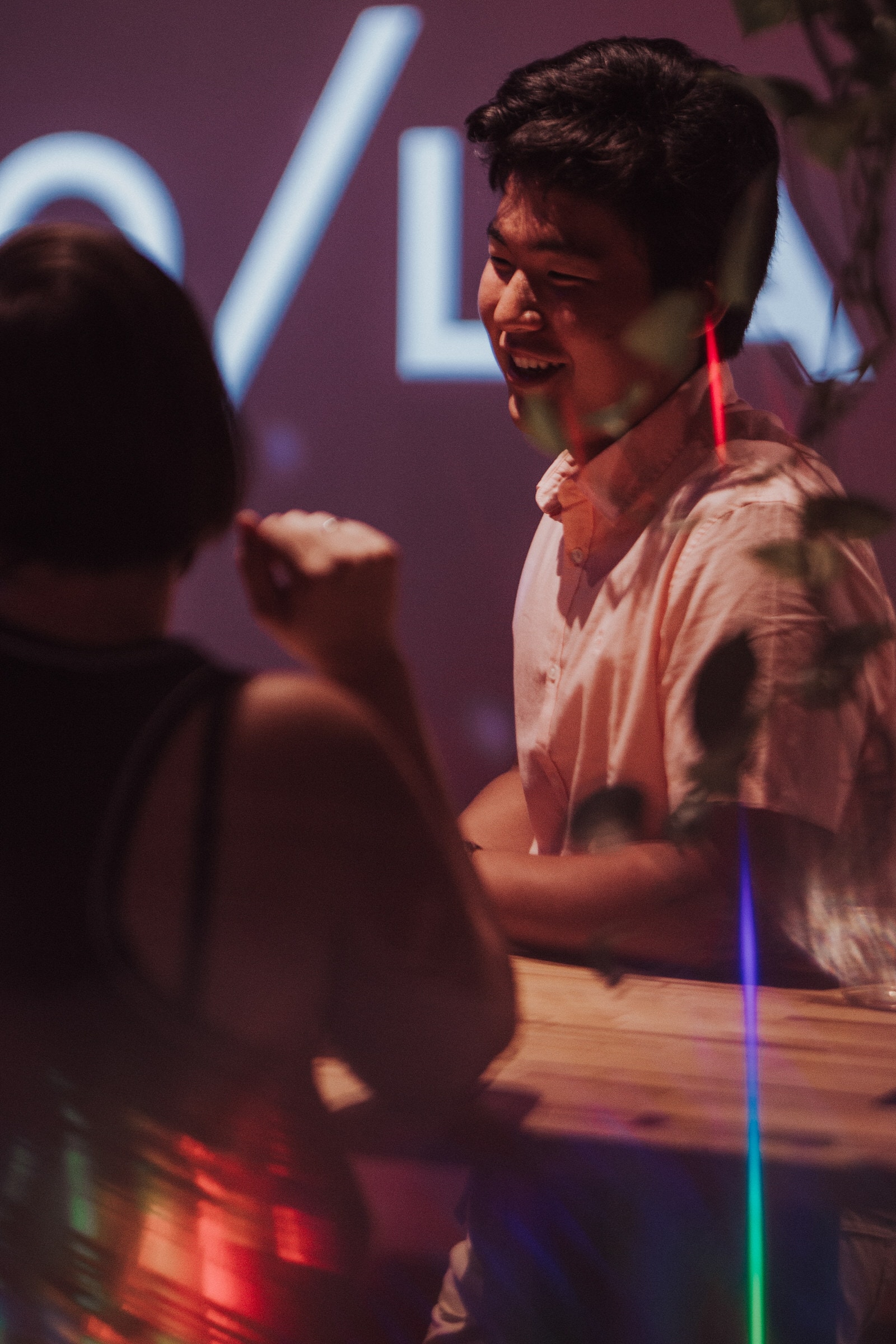 Man laughing while talking at a wooden table, lit by neon signage at a night event.