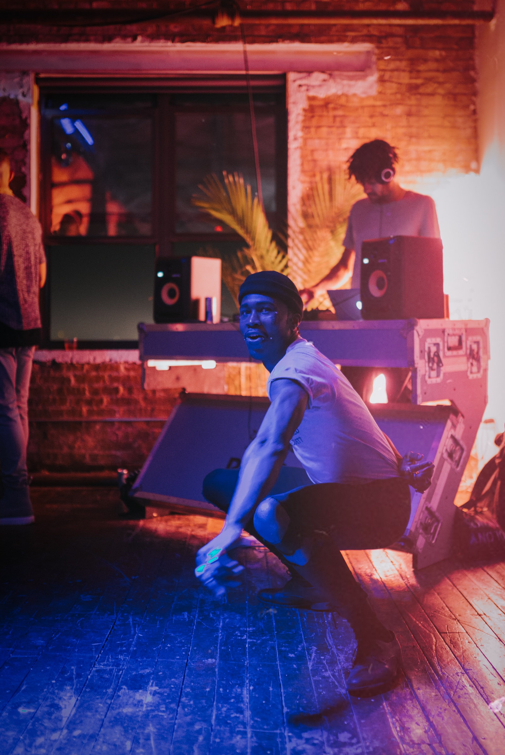 A man crouches on a dance floor as a DJ performs under blue and red club lighting.