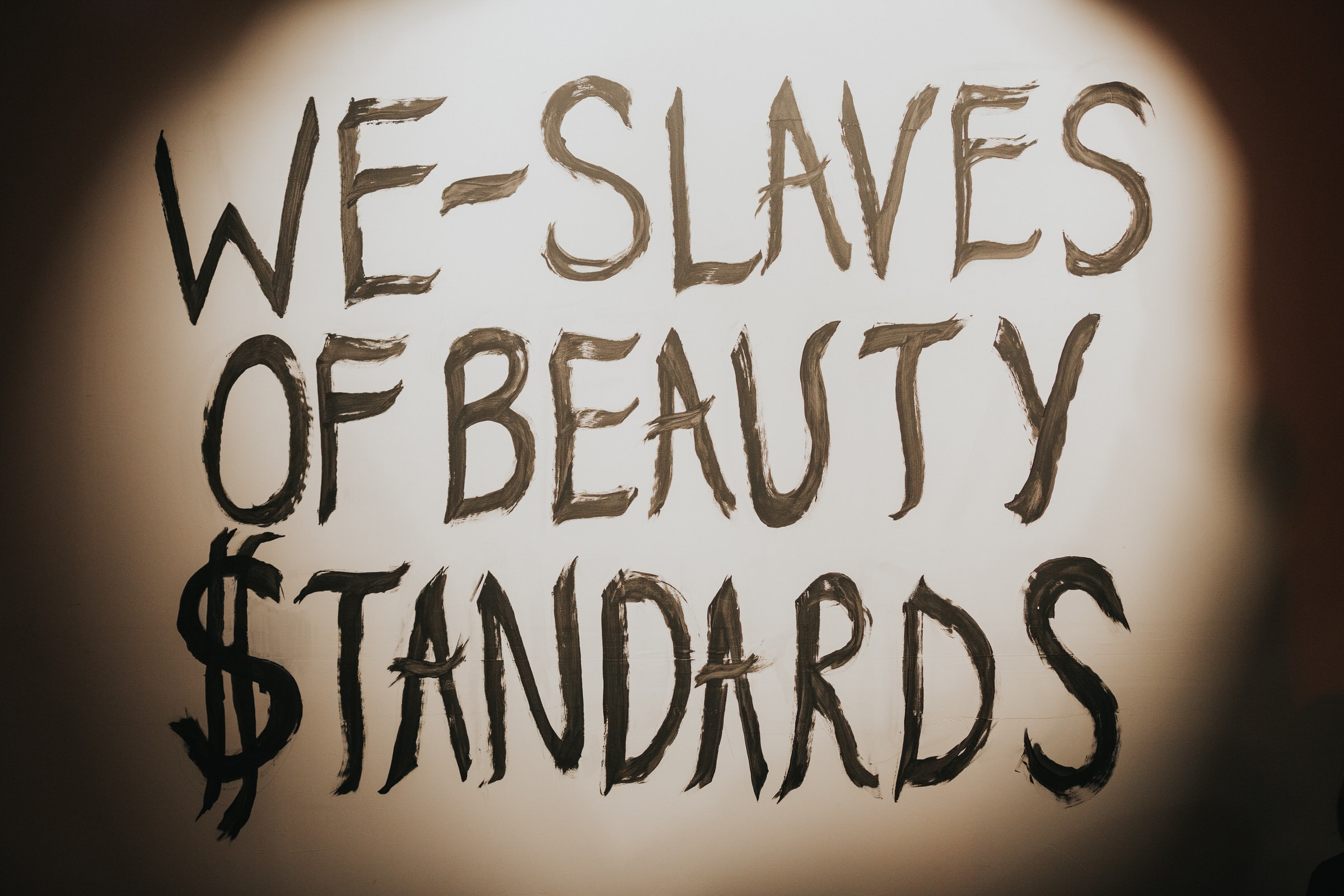 Hand-painted sign reading We Slaves of Beauty Standards, lit dramatically at an event.