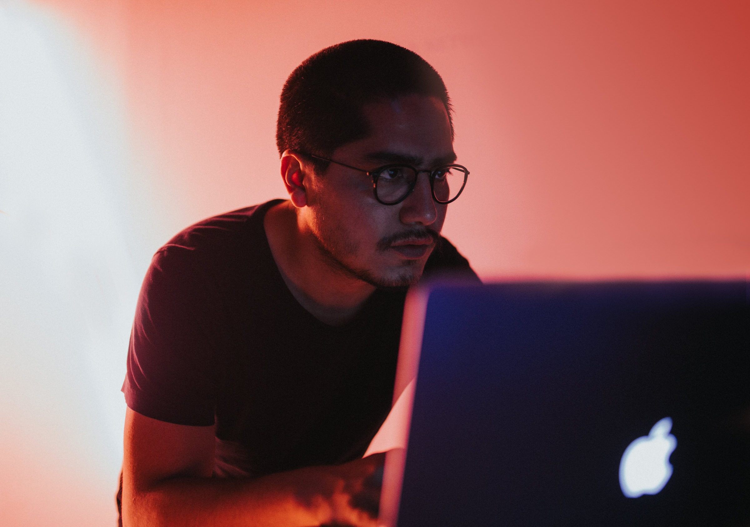 Man wearing glasses focuses on a MacBook screen under moody red studio lighting.