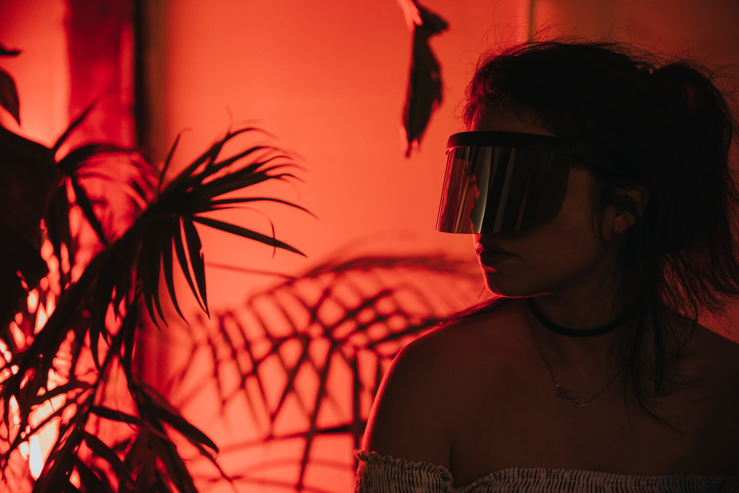 Woman in visor sunglasses stands beside palm leaves under red light at Colony Nights NYC.