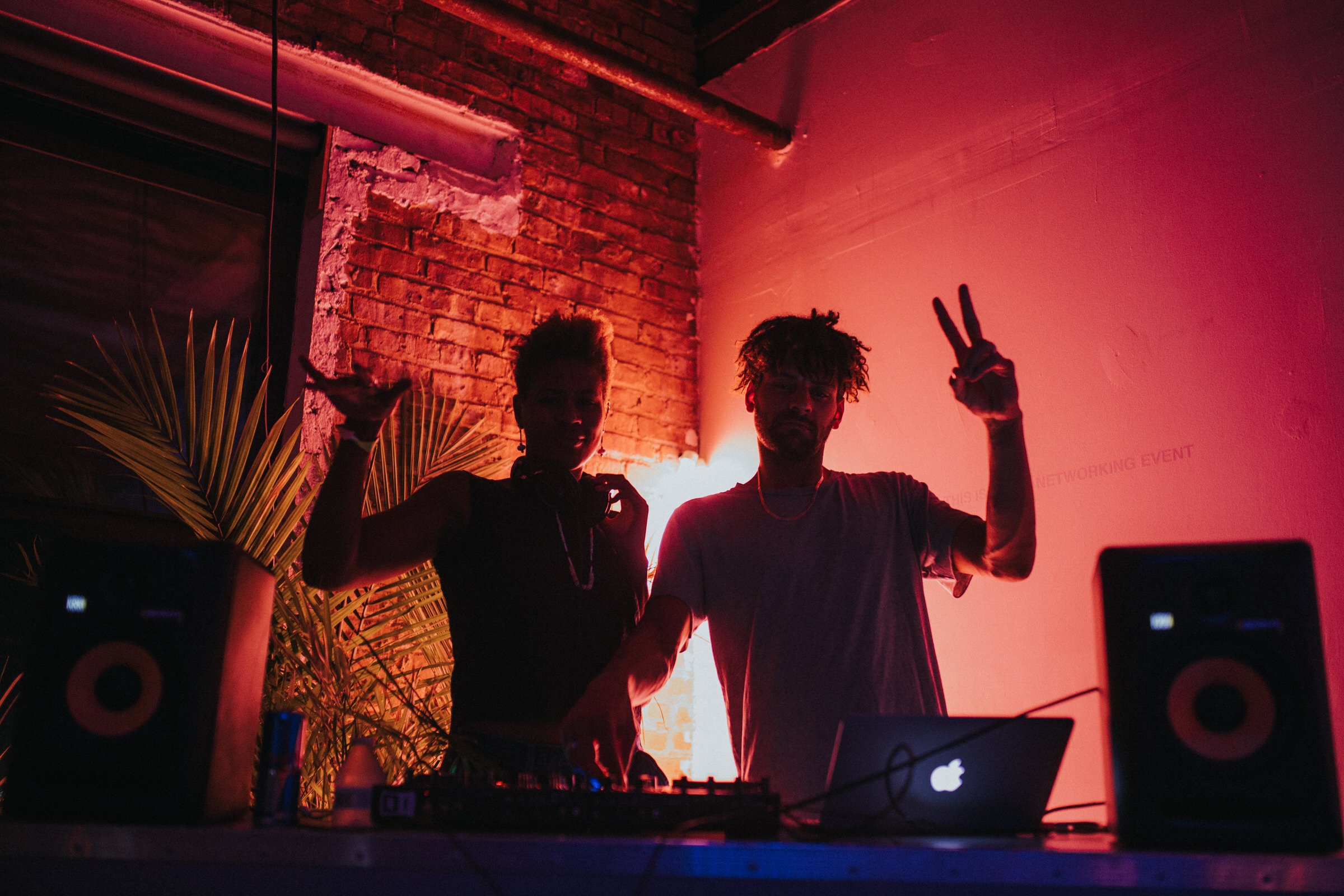 Two DJs gesture at the crowd behind a laptop and mixer under red club lighting.