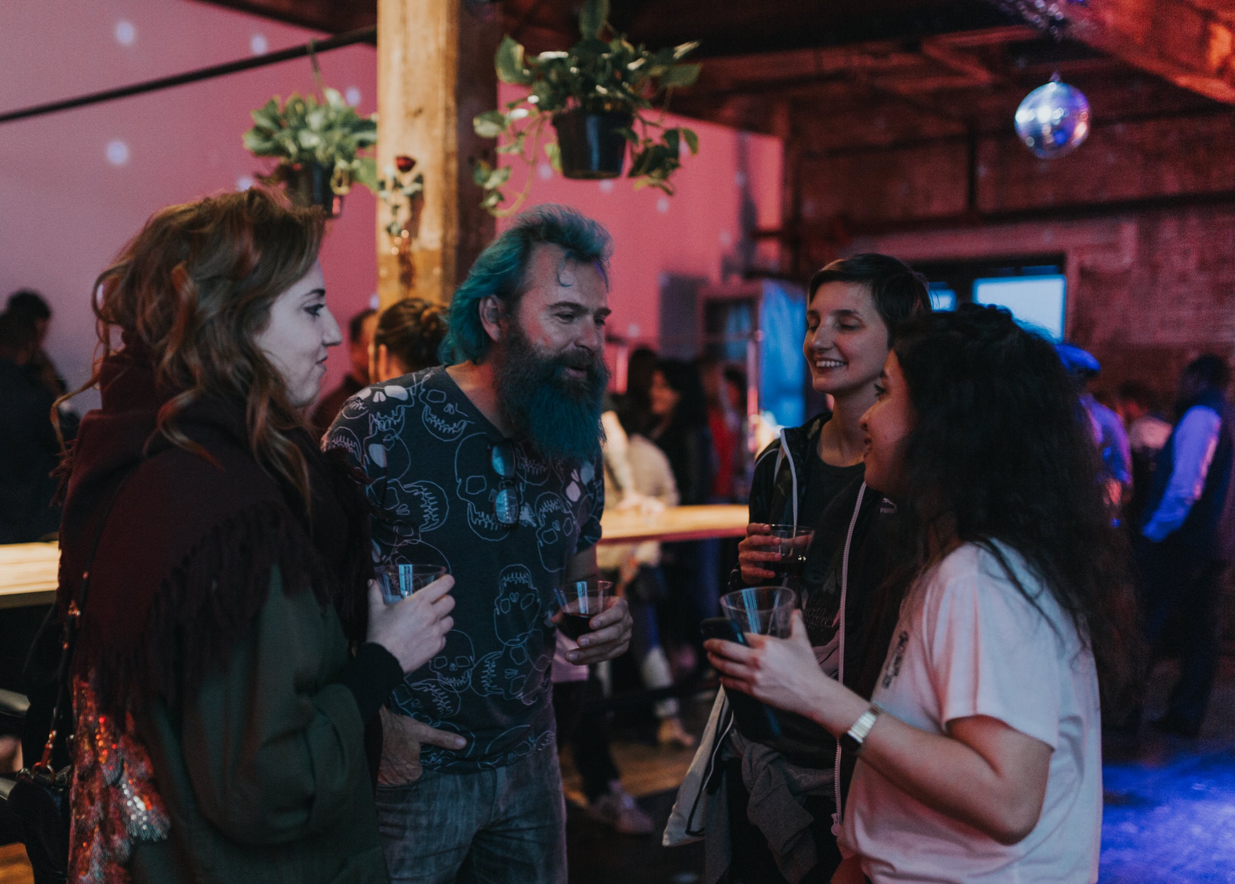 Guests chat with drinks at a dimly lit warehouse party under a disco ball.