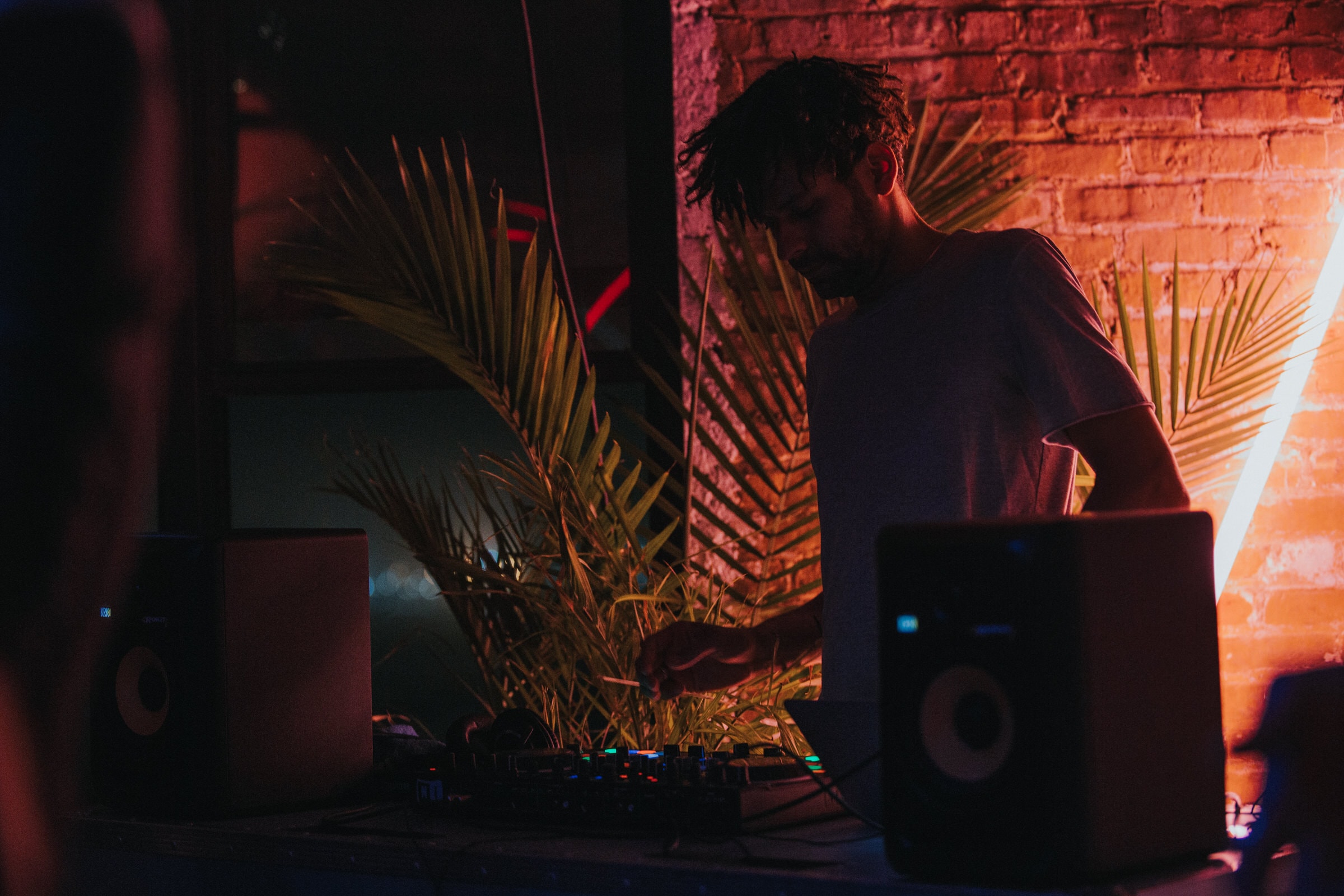 DJ mixing at a nighttime event, lit red beside brick wall and palm leaves.