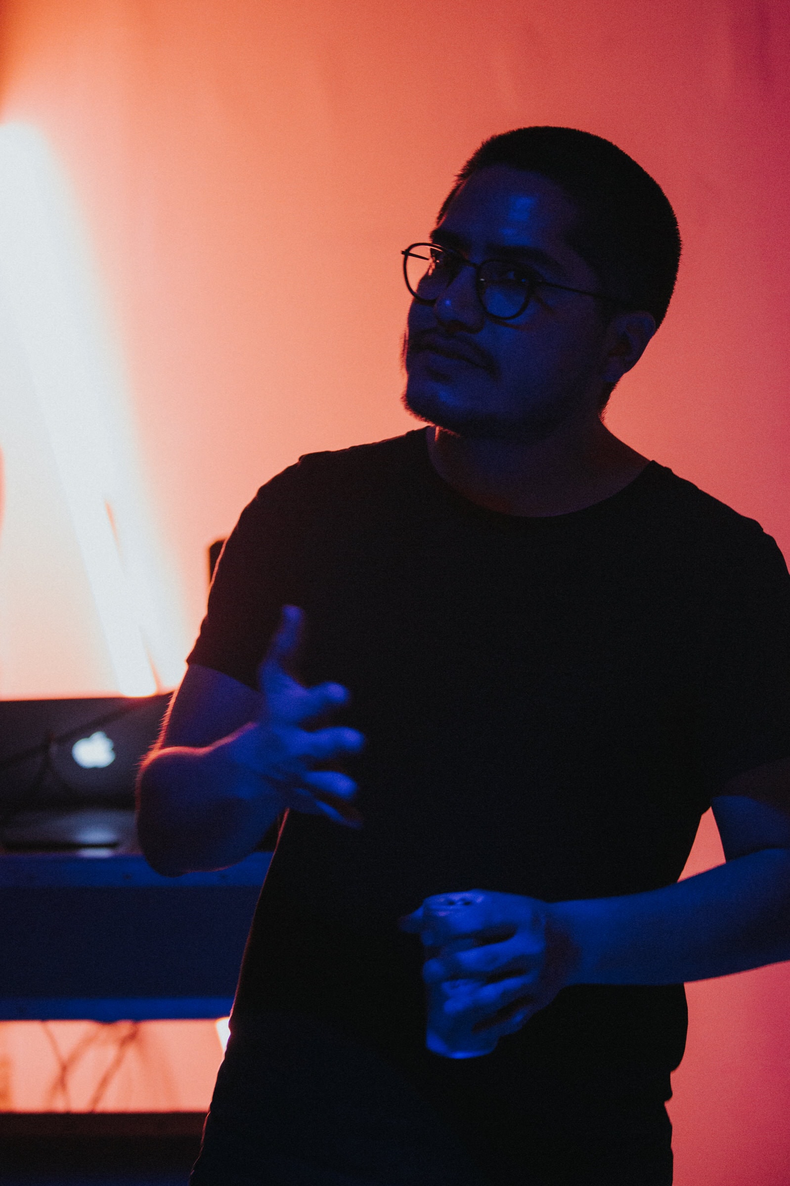 Man gestures while speaking at Colony Nights NYC, lit in blue and orange stage light.