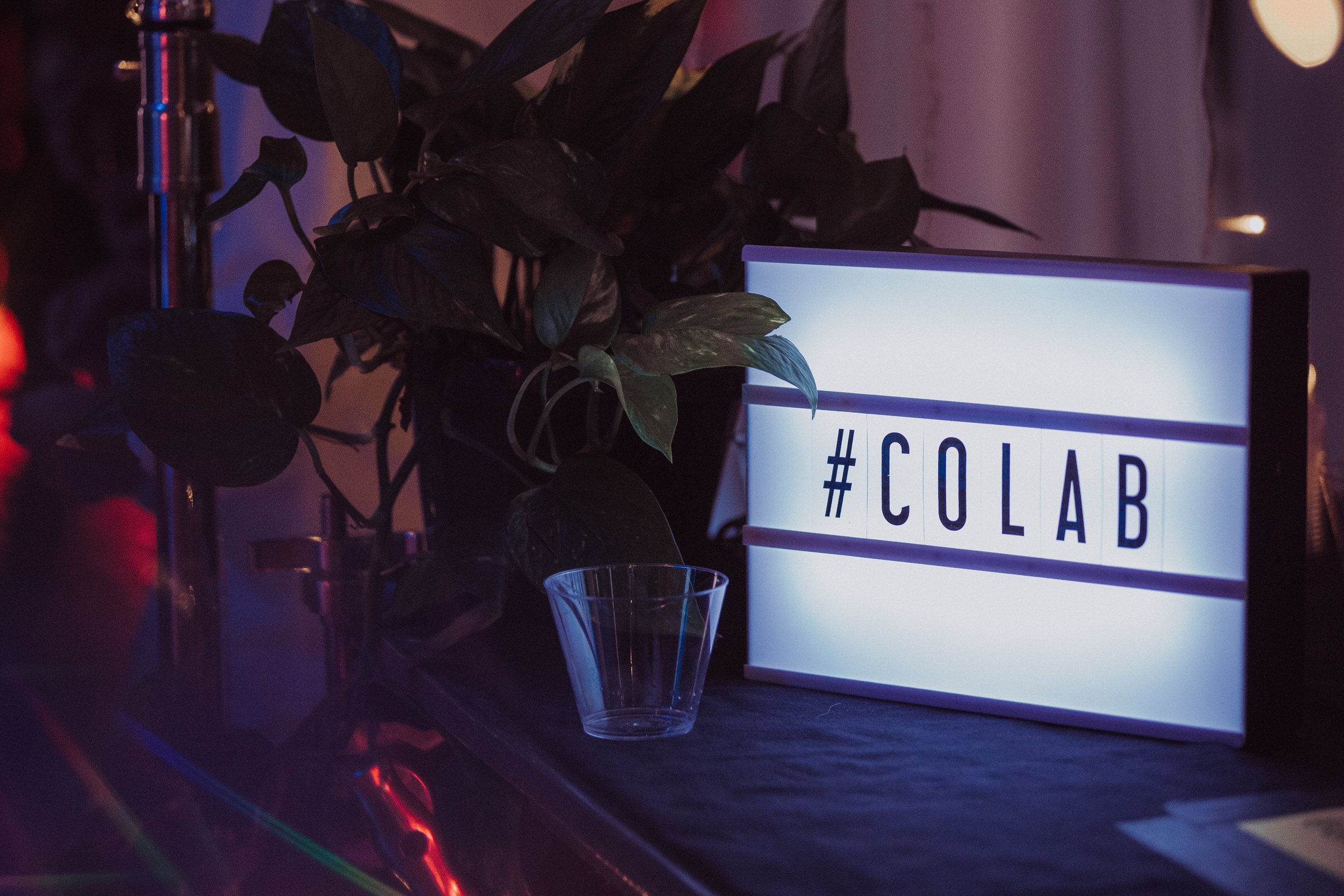 Lightbox sign reading hashtag colab beside a plant at a dim event venue.