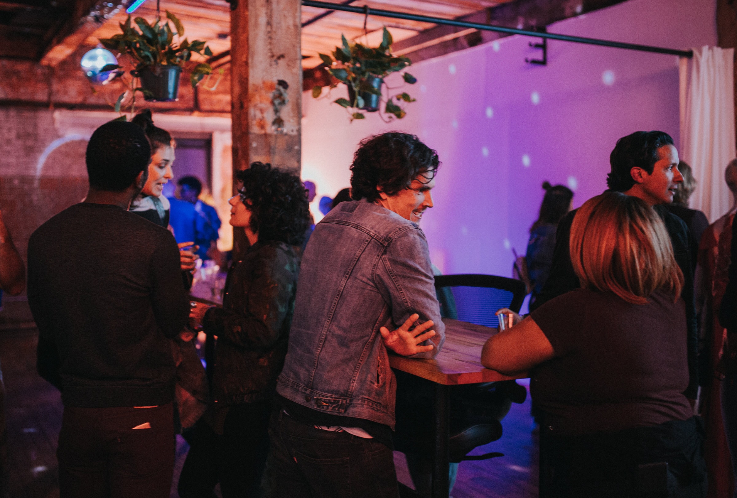 Guests mingle at a warehouse party under purple lighting and hanging plants.