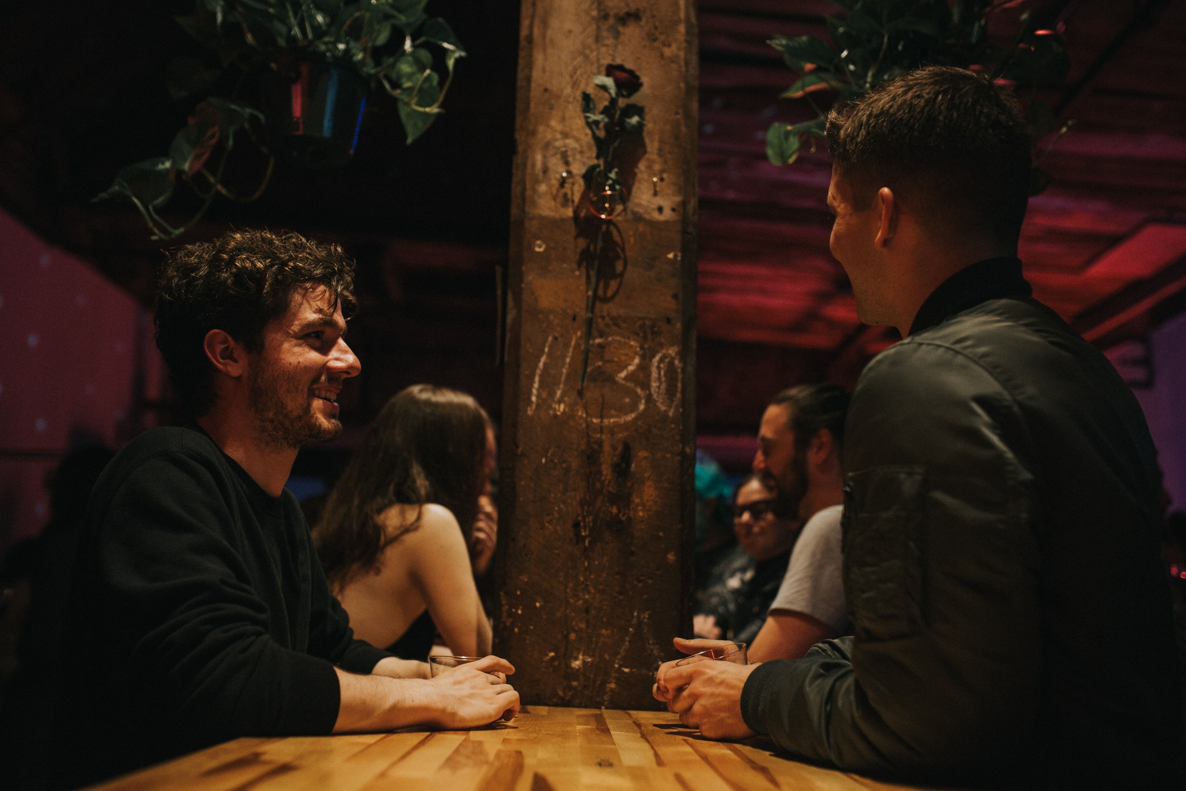 Two men chat over drinks at a wooden table in a dim, plant-filled bar.