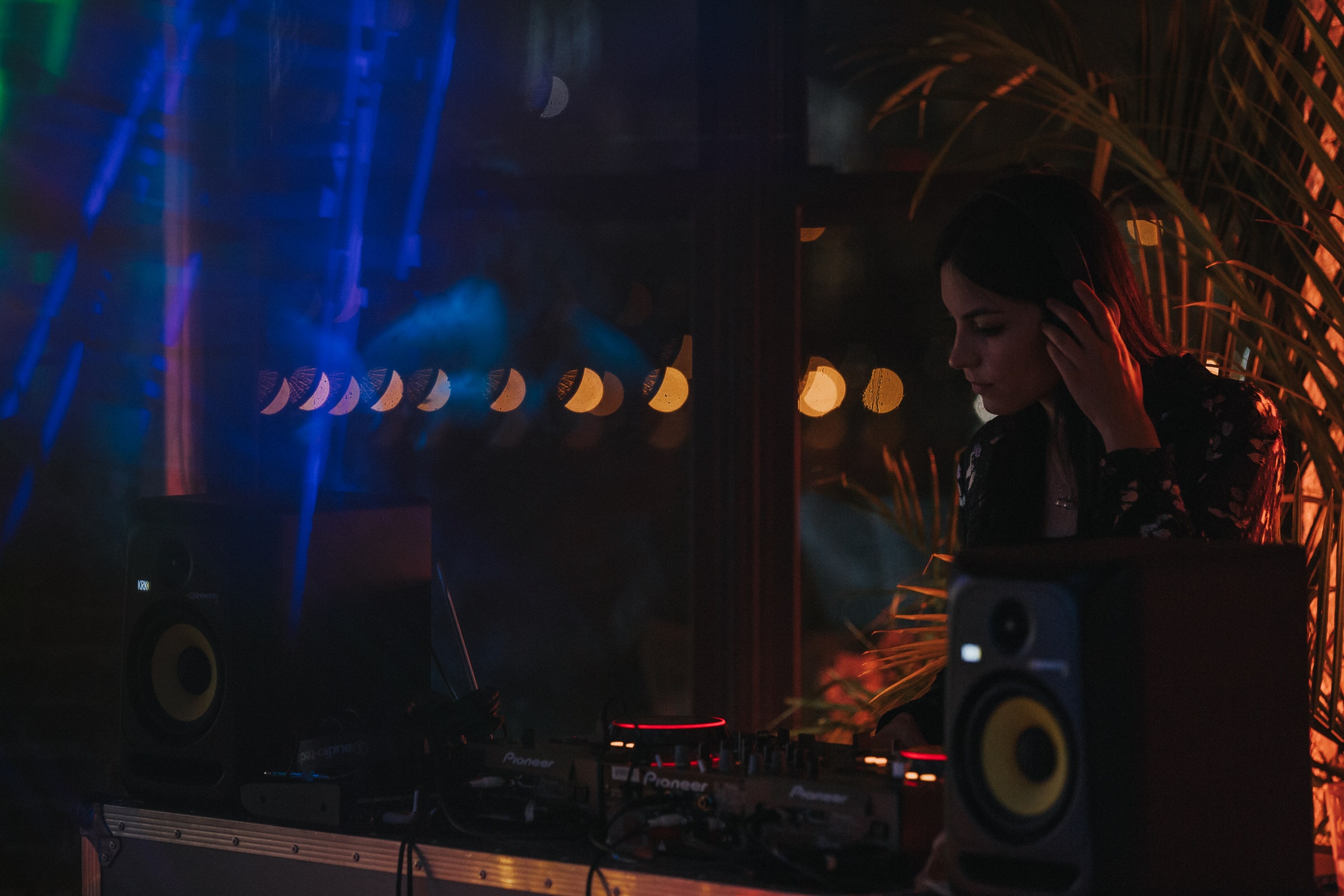 DJ performing at a dimly lit nightclub, surrounded by moon phase lights and palm fronds.