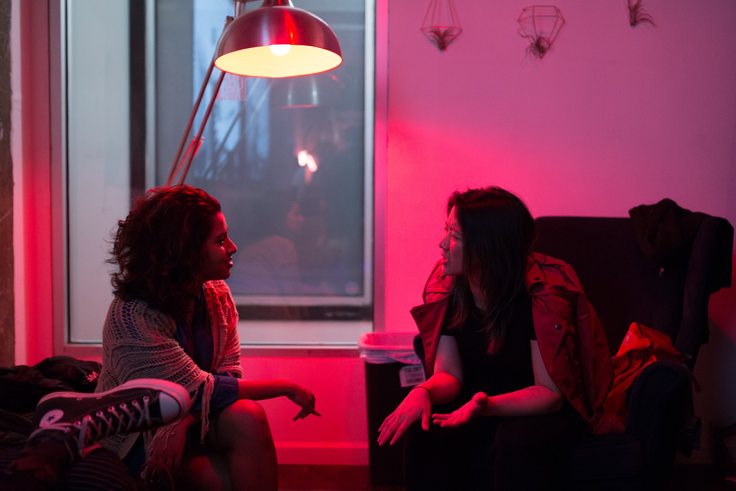 Two women talk under red lighting near a window at a nighttime event.
