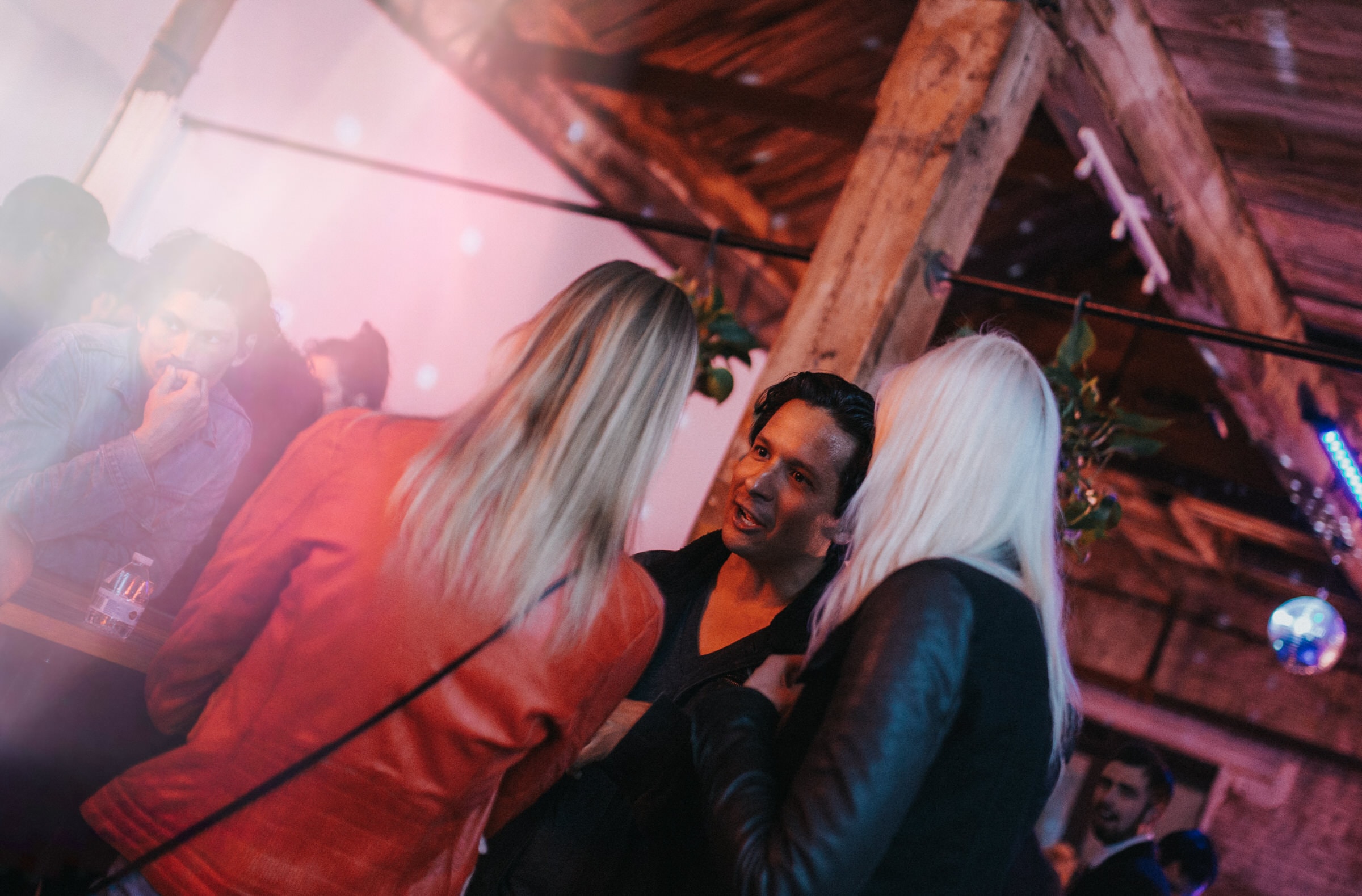 People talk closely under wooden rafters at a dimly lit nighttime event.