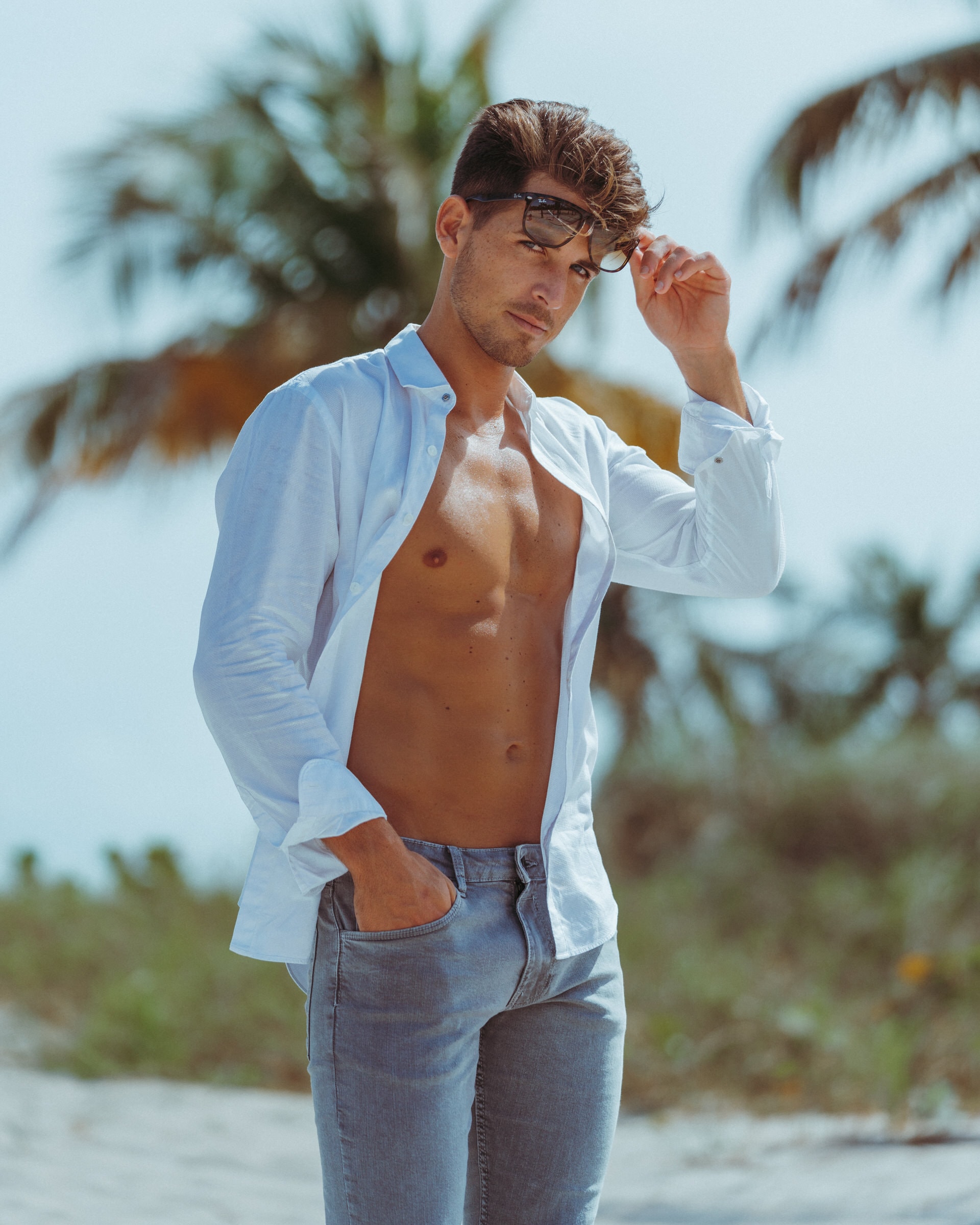 Man in an open white shirt adjusts sunglasses on a palm-lined beach.