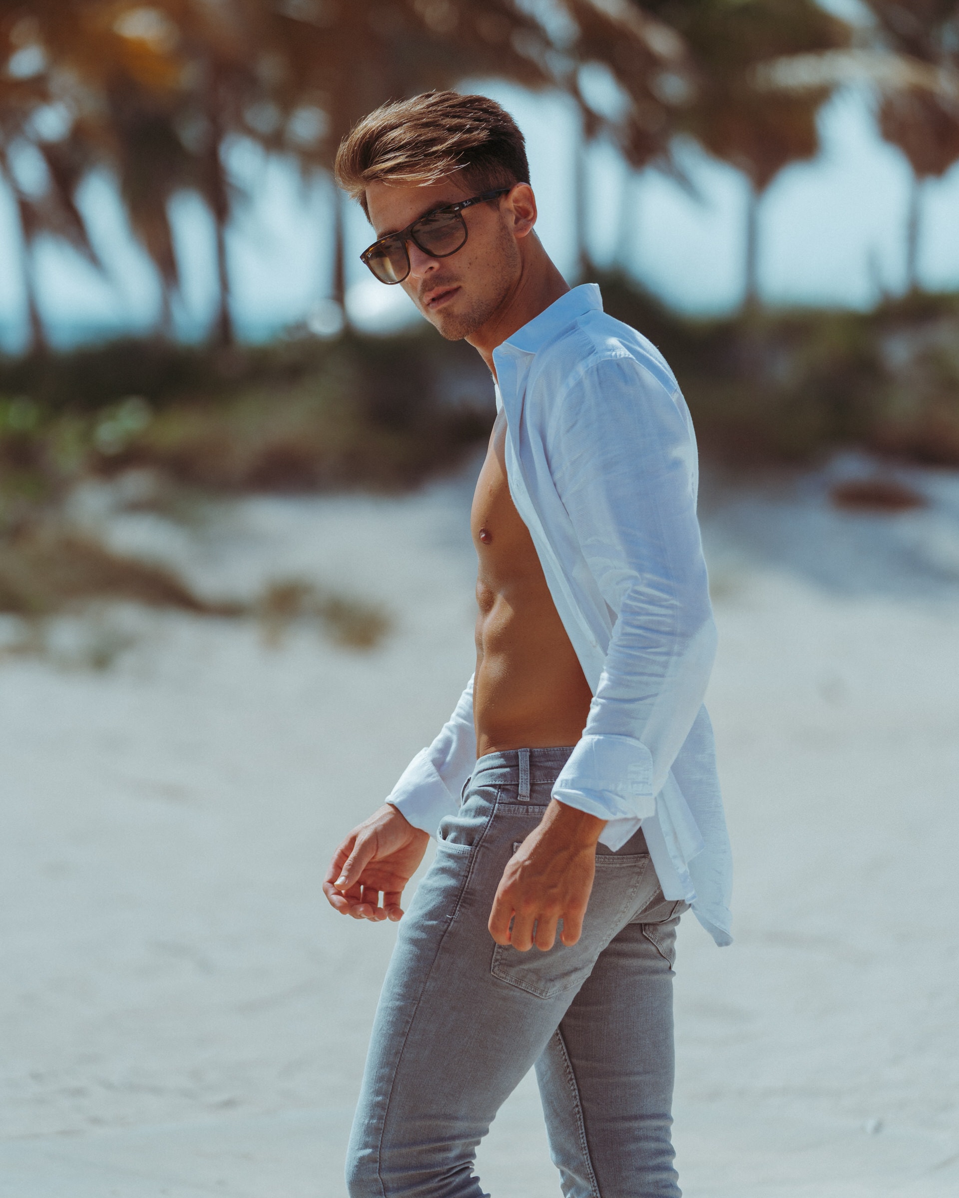 Man in open shirt and sunglasses poses on a sandy beach lined with palm trees.