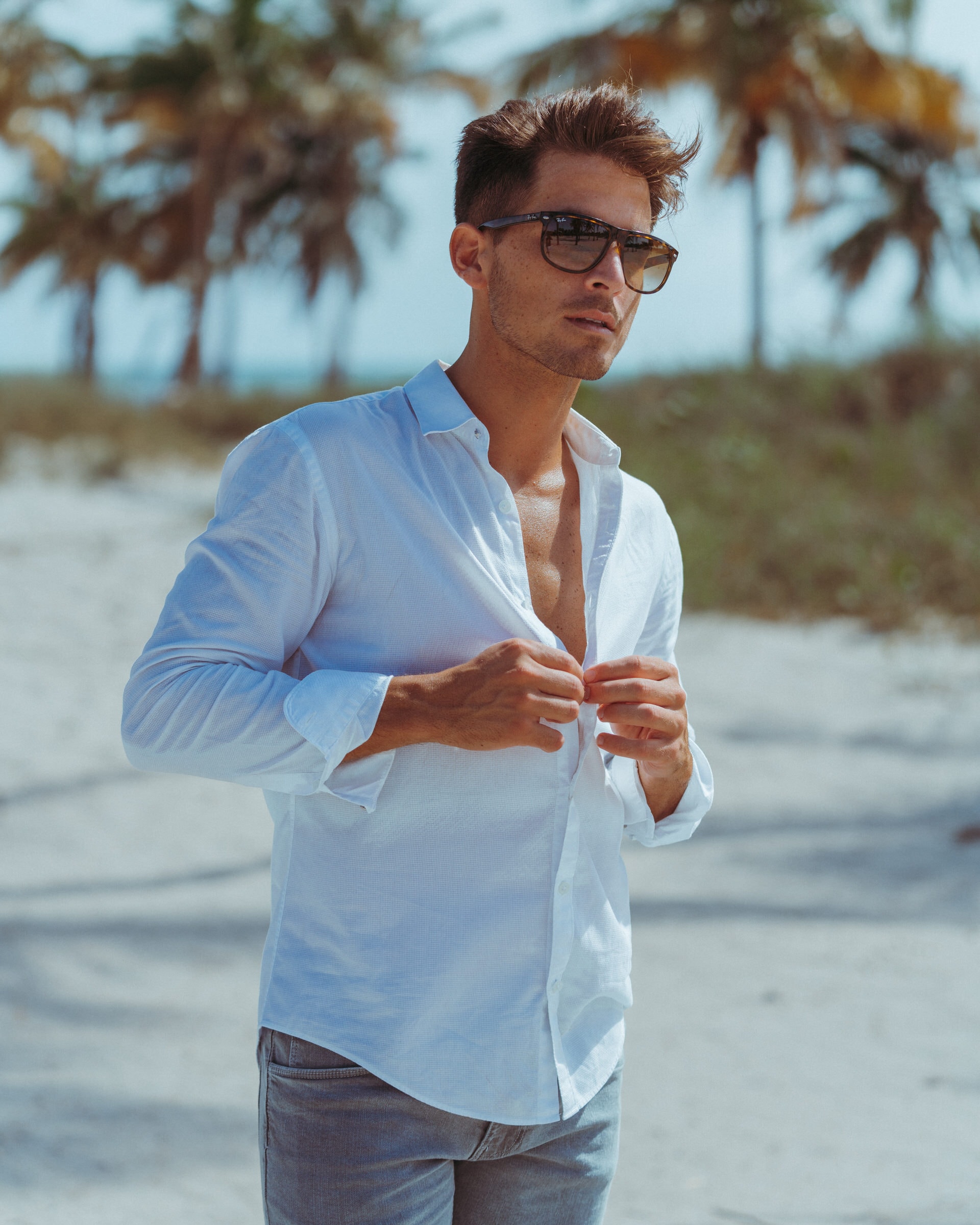 Man in sunglasses buttons a white linen shirt on a sandy beach with palm trees.