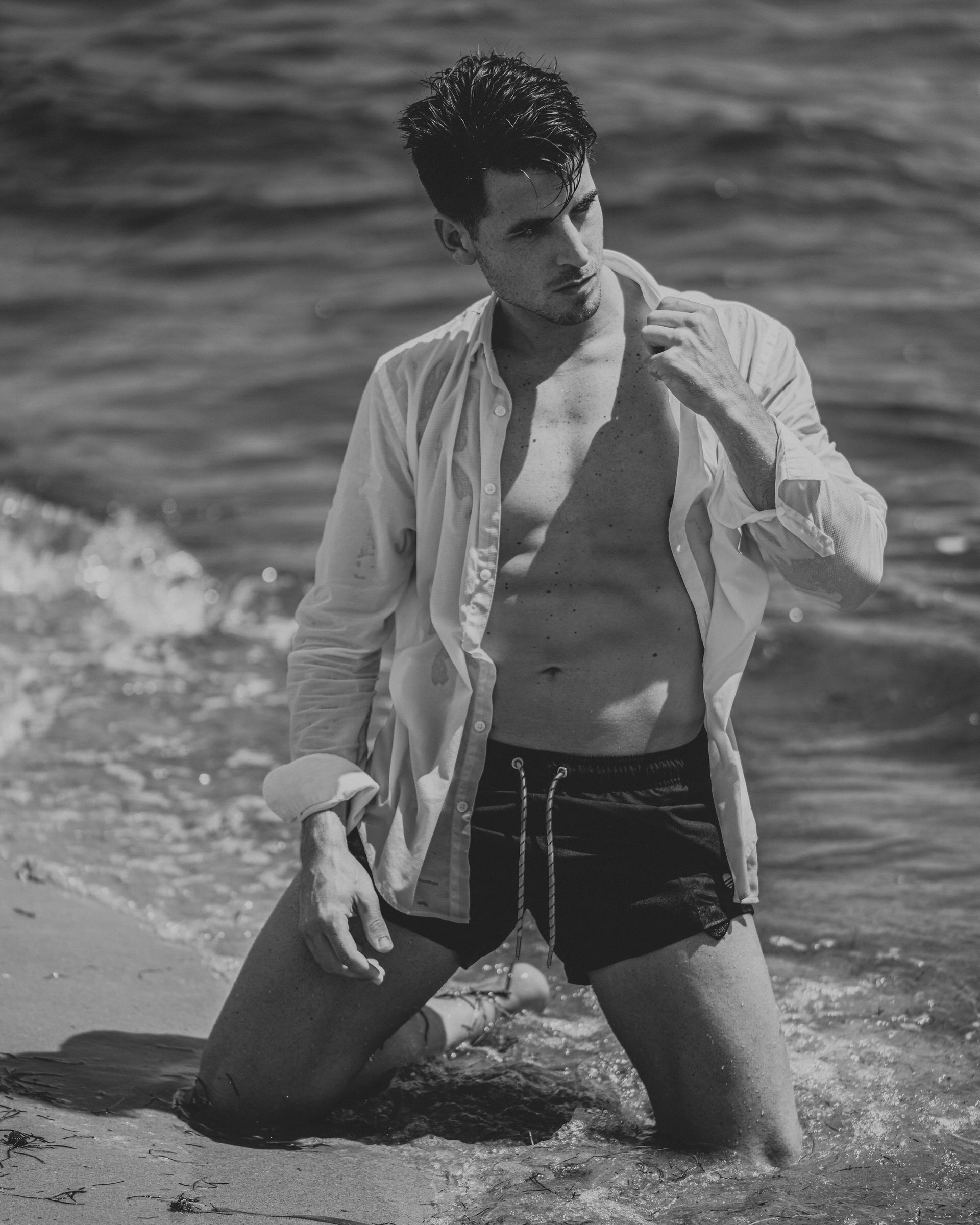Black and white beach portrait of a man kneeling in surf, wet shirt open.
